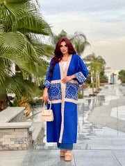 A woman wearing a blue cardigan with decorative patterns, paired with a white top and carrying a white bag, standing on a pathway with palm trees in the background.