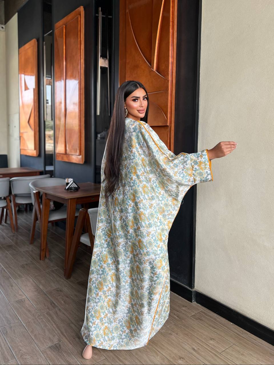 Woman in a floral dress standing in a room with wooden furniture and a mirror.